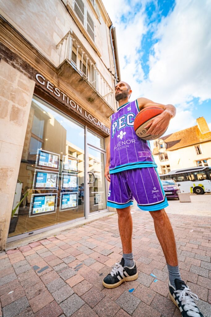Joueur du PEC Basket Poitiers, ballon en main, en maillot violet floqué Aliénor Gestion devant la vitrine de l'agence
